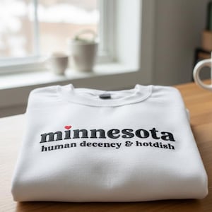 Pode incluir: Uma camisola branca dobrada com o texto bordado "minnesota human decency & hotdish" em preto. Um pequeno coração vermelho é bordado acima da palavra "minnesota". A camisola está numa superfície de madeira perto de uma janela.