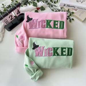 May include: Two sweatshirts with the word "WICKED" embroidered on them. One sweatshirt is pink and the other is green. Both sweatshirts have a small witch's hat embroidered above the word "WICKED".