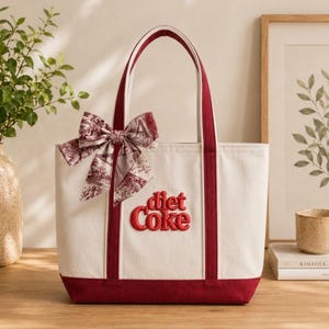 Może przedstawiać: Kremowa torba typu tote z płótna z ciemnoczerwonymi wykończeniami i uchwytami. Z przodu znajduje się wyhaftowany czerwony napis Diet Coke. Jedwabna apaszka we wzór toile jest zawiązana w kokardę wokół lewego uchwytu torby.