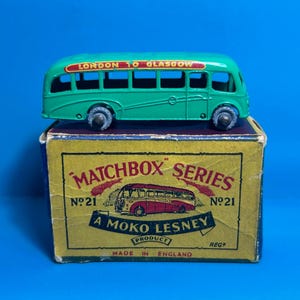 Puede incluir: Autobús de juguete verde con "LONDON TO GLASGOW" en el lateral, sentado en una caja amarilla con "MATCHBOX SERIES N°21 A MOKO LESNEY PRODUCT MADE IN ENGLAND REG" impreso en ella.