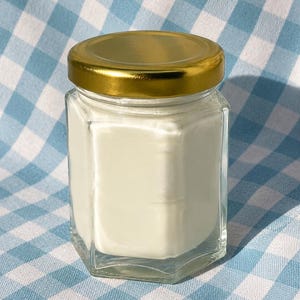 Könnte beinhalten: Ein klares, sechseckiges Glasgefäß, gefüllt mit einer glatten, undurchsichtigen weißen Substanz. Das Glas ist mit einem glänzenden, goldfarbenen Schraubdeckel aus Metall verschlossen. Der Behälter hat flache vertikale Seiten und einen stabilen Boden.