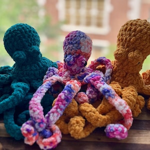 May include: Three crocheted octopus plush toys in teal, multicolored, and orange. The plushies have textured bodies and long, curled tentacles. The background is blurred, with a window and natural light.