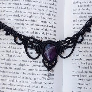 Peut inclure: Collier en macramé noir avec une grosse pierre précieuse violette en forme de goutte d'eau au centre. Le collier présente des nœuds complexes et de petites perles argentées. Un croissant de lune argenté pend en dessous, sur une page de livre.