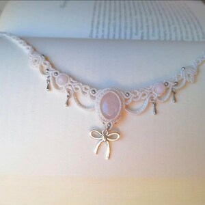 May include: A white macrame necklace with a central oval rose quartz stone and smaller rose quartz beads. The necklace has a silver bow pendant and small silver teardrop charms. The design is intricate and feminine.