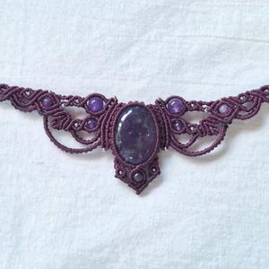 Puede incluir: Un collar de macramé artesanal en un tono púrpura intenso, con una pieza central de amatista ovalada y cuentas de amatista más pequeñas. El intrincado diseño de nudos está acentuado con pequeñas cuentas de color plateado, creando una joya única y elegante.