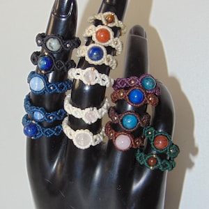 Può includere: Una collezione di anelli in macramè fatti a mano esposti su un porta gioielli nero a forma di mano. Gli anelli presentano pietre preziose di vari colori, tra cui blu, rosa e arancione, incastonate in intricati motivi intrecciati. Gli anelli sono in vari colori, tra cui bianco, blu e marrone.