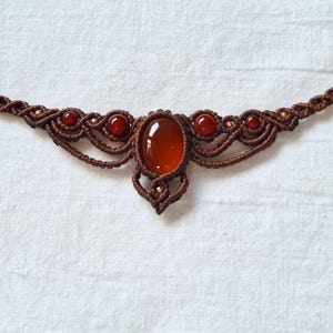 Peut inclure: Un collier en macramé marron orné d'une pierre centrale ovale rouge-orangé et de petites pierres rondes. Le design tissé complexe comprend des perles dorées, créant un style bohème et artisanal.