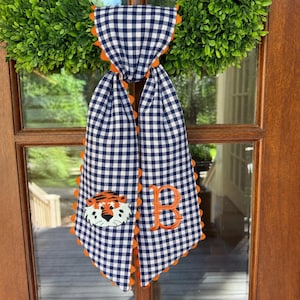 May include: A navy blue and white gingham fabric door hanger featuring a scalloped orange trim. The left side displays an embroidered tiger face, while the right side features an embroidered orange letter B in a serif font style.
