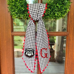 May include: A black and white gingham fabric scarf featuring a red scalloped edge trim. One side displays an embroidered bulldog head graphic, while the other side features a large red embroidered letter G. The scarf hangs vertically.
