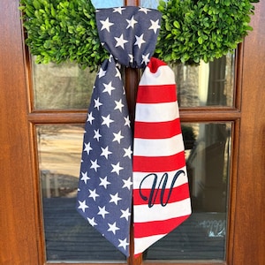 May include: A patriotic door hanger featuring a green wreath with a blue fabric section adorned with white stars and a red and white striped section with a black initial "W". The door decoration is designed to hang on a door.