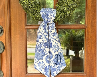 May include: A blue and white floral patterned scarf with the letter "D" embroidered on it. The scarf is hanging on a wooden door with a green wreath in the background. The scarf's design features blue flowers and leaves.