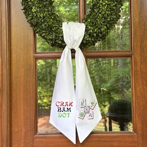 May include: White fabric door decoration with embroidered text and Mahjong tiles. The text reads "Crak Bam Dot" in red, black, and green. The Mahjong tiles are green, red, and white. A green wreath hangs above the decoration.