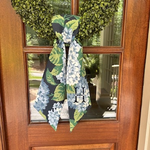 以下が含まれることがあります： 青、緑、白の花柄模様の装飾ドアスカーフ。文字「H」が特徴です。 スカーフは緑のリースに結ばれており、ドアにエレガントさを加えています。 スカーフは柔らかく流れるような生地で作られています。