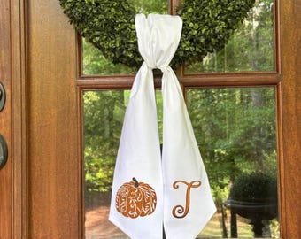 Puede incluir: Faja de puerta blanca con una calabaza naranja decorativa y la letra "J" impresa. La faja cuelga de una puerta de madera con una corona verde. El diseño de la calabaza tiene detalles intrincados.