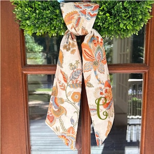 May include: A long, narrow fabric scarf featuring a beige background with a repeating floral pattern in shades of orange, blue, and green. The scarf is tied in a knot and displays a large, embroidered olive green letter C on the lower right side.
