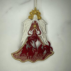 May include: Embroidered Christmas ornament depicting an angel with white wings and a gold halo. The angel's red and gold robe shows three camels and palm trees. The ornament is suspended by a gold string.