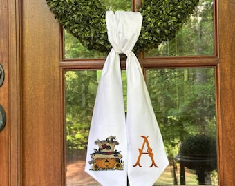 Puede incluir: Un colgador de puerta de tela blanca drapeado sobre una corona de boj verde. El lado izquierdo presenta una calabaza bordada colorida con un sombrero de girasoles, mientras que el lado derecho muestra una letra A naranja bordada en fuente serif.