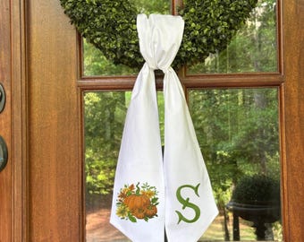 Puede incluir: Adorno de puerta blanco con una "S" verde y un diseño de calabaza y flores. El adorno cuelga de una puerta de madera con una corona verde. El diseño presenta calabazas naranjas y hojas amarillas y verdes.