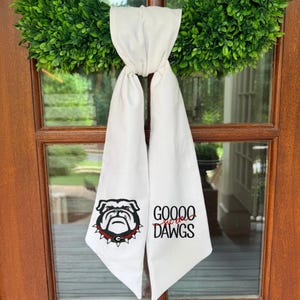 May include: A white fabric neck scarf featuring a black and red bulldog mascot graphic on one side and the black text GOOOO DAWGS with red script text that reads sic em on the other side. The scarf is tied in a knot at the top center.