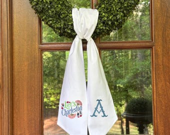May include: White fabric door sash with the text "Let's Charleston" and the letter "A" embroidered in blue. The sash is tied to a green wreath on a wooden door.