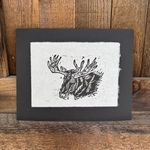 May include: A black and white linocut print depicting a moose with prominent antlers. The artwork is presented on textured, off-white paper, framed by a black mount. The print is set against a wooden backdrop.