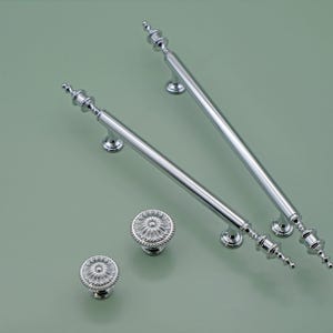 Może przedstawiać: Dwa srebrne uchwyty do szafek z ozdobnymi zakończeniami i dwa pasujące srebrne gałki z kwiatowym wzorem.