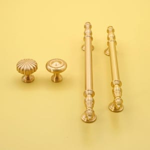 Può includere: Una collezione di ferramenta per mobili in ottone, tra cui due pomelli e due maniglie. I pomelli presentano dettagli decorativi, e le maniglie sono cilindriche con estremità ornate. La ferramenta è posta su uno sfondo giallo.