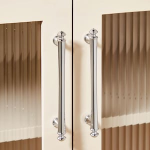 以下が含まれることがあります： クリーム色のキャビネットに、ガラスパネルと装飾的なフィニアルが付いたクロム製のキャビネットドアハンドルが2つあります。