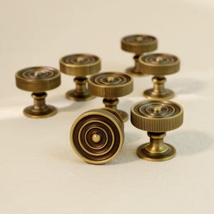 May include: A collection of antique brass cabinet knobs. Each knob features a circular design with concentric rings and a ridged edge. The knobs are mounted on a short, rounded base, and are a warm, golden color.