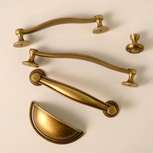 May include: An assortment of antique brass cabinet hardware, including curved handles, a knob, and a cup pull. The hardware has a warm, aged finish and decorative detailing, perfect for kitchen or bathroom renovations.