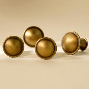 May include: A collection of five antique brass cabinet knobs. Each knob has a rounded, domed shape with a slightly textured surface and a warm, golden-bronze color. The knobs are arranged on a neutral background.