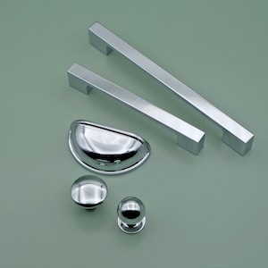 Könnte beinhalten: Ein Satz aus fünf verchromten Schrankbeschlägen, darunter zwei lange Griffe, ein Tassen-Griff und zwei Knöpfe. Die Beschläge sind auf einer hellgrünen Oberfläche angeordnet.