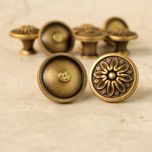 Könnte beinhalten: Eine Sammlung antiker Messing-Schrankknöpfe. Die Knöpfe weisen detaillierte Designs auf, darunter ein Blumenmuster und eine kleine Blume. Die Knöpfe haben ein warmes, gealtertes Aussehen, das auf Vintage- oder Retro-Stil schließen lässt. Die Knöpfe sind rund und haben einen Stiel.