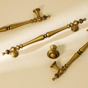 May include: A selection of antique brass cabinet handles and a knob. The handles feature detailed designs and a warm, golden colour. The hardware is displayed on a neutral background, highlighting the vintage style. The handles are of varying lengths, suitable for different cabinet sizes.