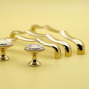 Puede incluir: Cuatro tiradores de armario de color dorado con un diseño ondulado y dos pomos dorados a juego con un patrón floral decorativo.