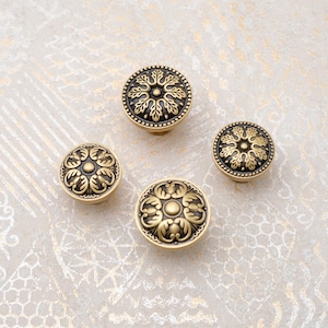 May include: Four antique brass cabinet knobs with intricate floral designs. The knobs feature a dark, almost black, center with a detailed pattern, surrounded by a lighter brass border. The knobs vary slightly in size, showcasing a classic hardware aesthetic.