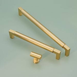 Può includere: Tre maniglie per armadi dorate e un pomello dorato. Le maniglie sono rettangolari con bordi arrotondati e sono disponibili in diverse dimensioni. Il pomello è rotondo con una base quadrata.