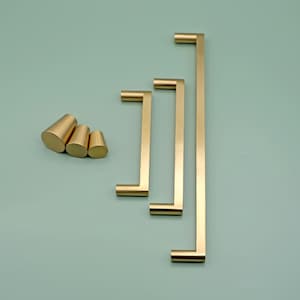 May include: A collection of gold-colored cabinet hardware, including three rectangular bar pulls of varying lengths and three conical knobs. The hardware is displayed against a muted green background, showcasing a modern design aesthetic.