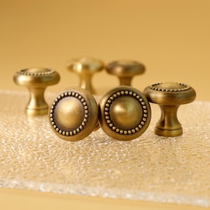 Può includere: Primo piano di sei pomelli per mobili in ottone antico. I pomelli hanno un design rotondo e bordato intorno al centro. I pomelli sono su una superficie trasparente e strutturata con uno sfondo giallo. I pomelli sono di un caldo colore ottone invecchiato.