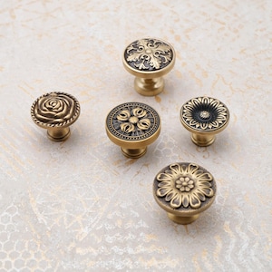 Könnte beinhalten: Fünf antike Messing-Schrankknöpfe mit aufwendigen Blumen- und Scroll-Designs. Die Knöpfe variieren in Form und Muster, darunter eine Rose, eine Blume und verzierte kreisförmige Motive. Jeder Knopf hat einen kurzen, abgerundeten Sockel.