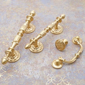 Puede incluir: Un conjunto de herrajes dorados y ornamentados para armarios, que incluye dos tiradores largos, un pequeño pomo y un tirador curvo. Los herrajes presentan intrincados diseños florales y de volutas, perfectos para añadir un toque de elegancia a los muebles.
