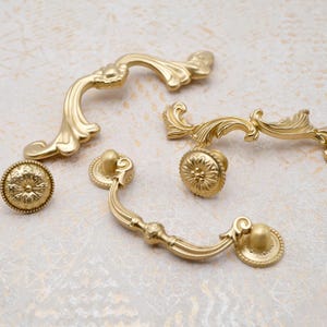 May include: A collection of gold-colored cabinet hardware, including ornate handles and knobs. The handles have a curved design with decorative flourishes, while the knobs feature a floral pattern. The hardware is arranged on a light-colored, textured surface.