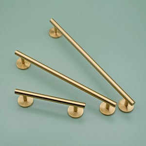 May include: Three gold-toned cabinet pulls with a cylindrical shape and a circular base. The pulls are arranged in a staggered pattern on a light blue background.
