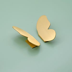 Pode incluir: Duas puxadores de armário dourados em forma de borboleta. Um puxador tem formato de asa única e o outro de asa dupla. Os puxadores são feitos de metal escovado e estão sobre um fundo verde-azulado claro.