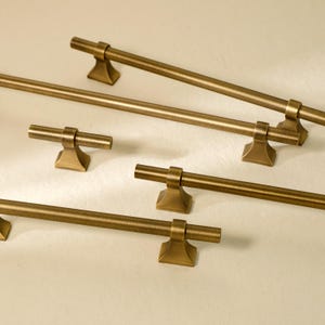Puede incluir: Varios tiradores y manijas de gabinete de color bronce están dispuestos sobre una superficie clara. El hardware presenta un diseño en forma de T con monturas de base cuadrada. Las manijas varían en longitud, lo que sugiere diferentes tamaños de gabinete.