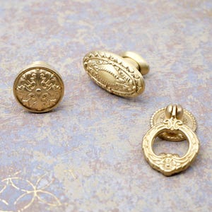 May include: Three antique brass cabinet hardware pieces. The hardware includes a round knob with a floral design, an oval knob, and a ring pull. The hardware is made of brass and has a gold finish.