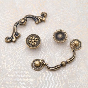 Könnte beinhalten: Eine Auswahl an antiken Messing-Schrankbeschlägen, darunter zwei Griffe und zwei Knöpfe. Die Griffe weisen verzierte Blumen- und Scroll-Designs auf. Die Knöpfe haben detaillierte Blumenmuster. Die Hardware hat ein Vintage-Aussehen.