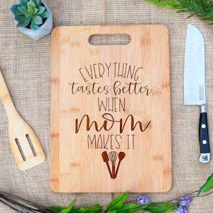 May include: A brown bamboo cutting board with the text "Everything tastes better when mom makes it" engraved on it. There are three kitchen utensils engraved below the text.