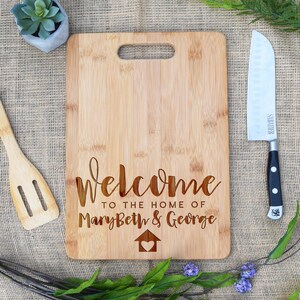 May include: A brown bamboo cutting board with a handle and the text "Welcome to the home of MaryBeth & George" engraved on it. The board has a small house with a heart inside engraved on it.