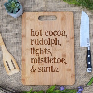 May include: A brown bamboo cutting board with the text "hot cocoa, rudolph, lights, mistletoe, & santa." engraved on it.